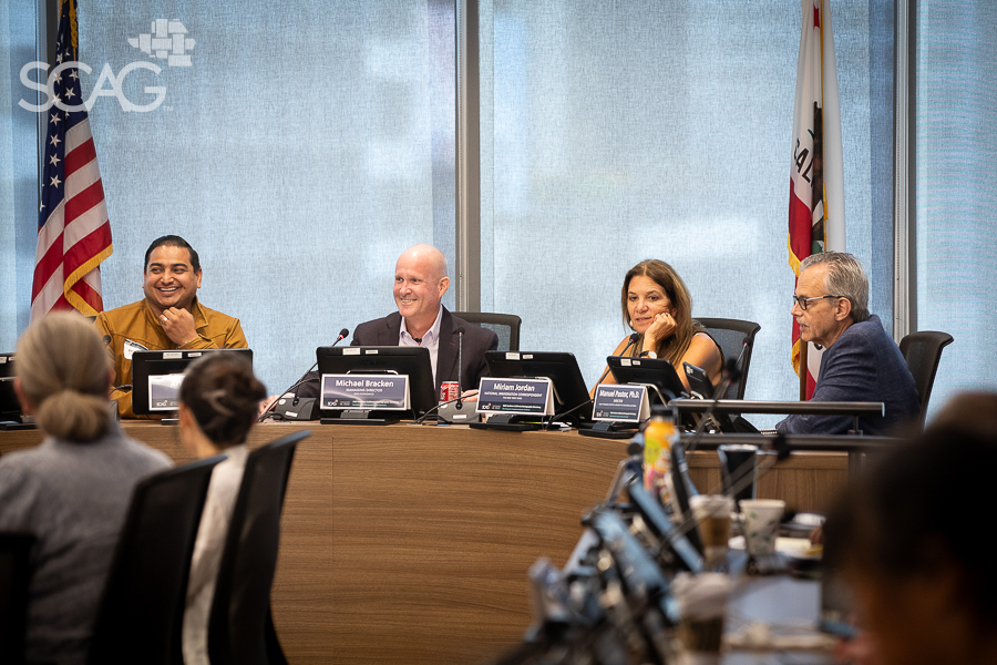 Four people seated at a conference table, engaged in discussion. U.S. and state flags behind them.
